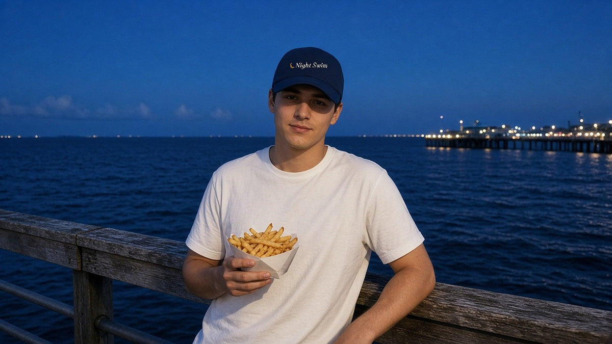 The Night Swim Dad Hat in Navy, worn at a boardwalk railing at blue hour with a boat of fries