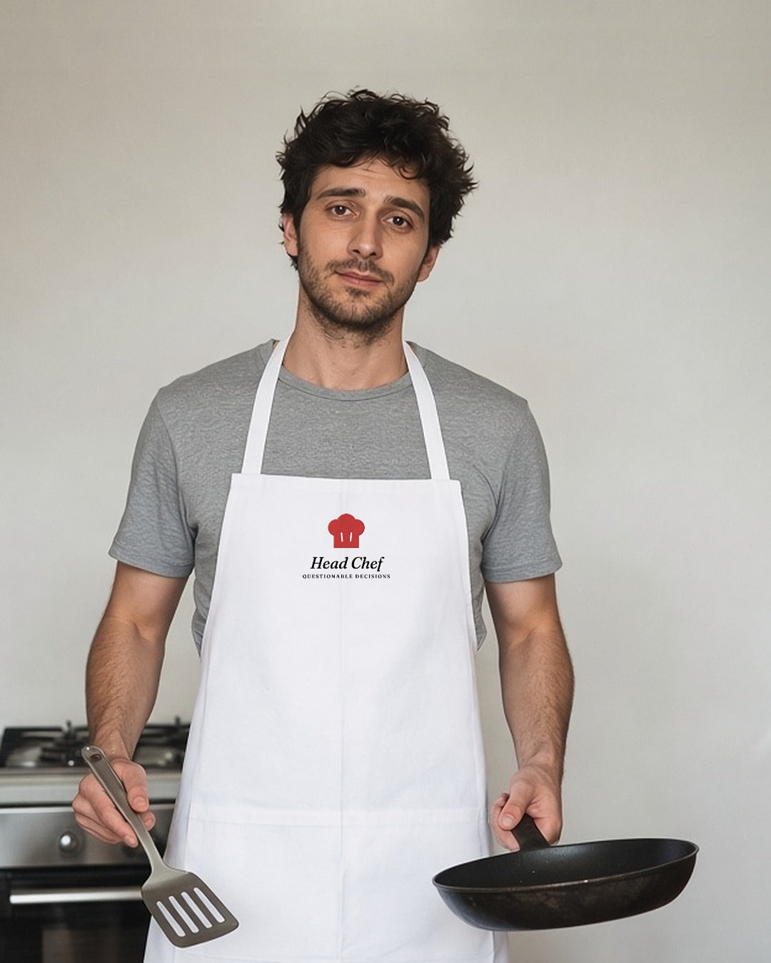 The Head Chef Apron in White, man in a kitchen holding a frying pan and a wooden spatula