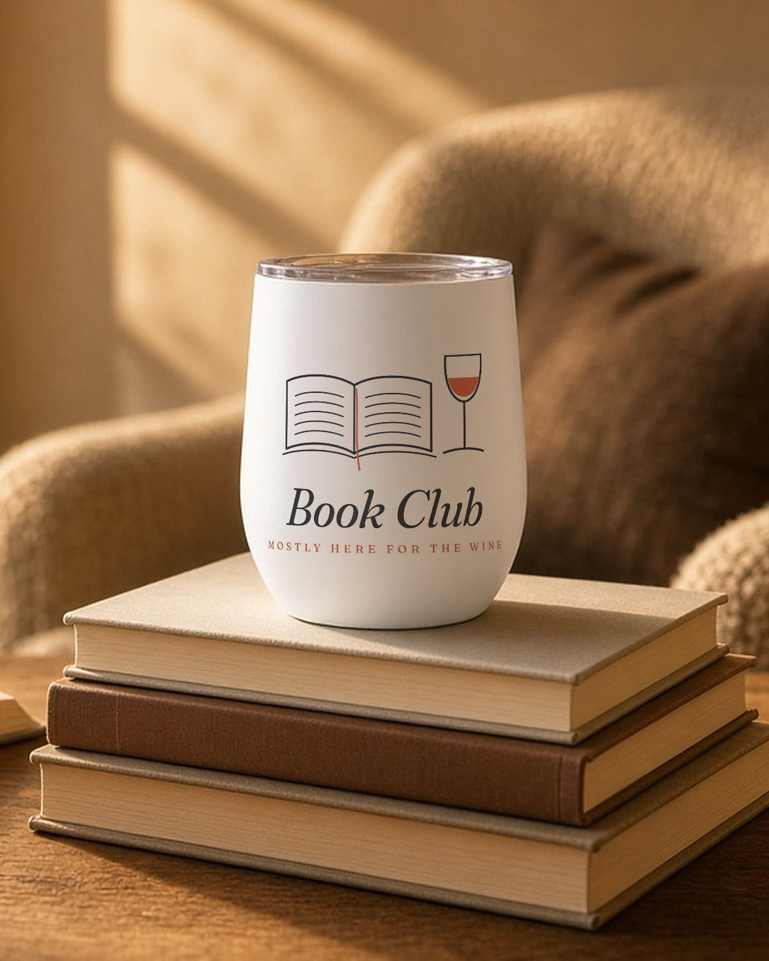 The Book Club Wine Tumbler in White, tumbler on a stack of books by an armchair