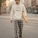 The Boulangerie Hoodie in Bone, a man (late 20s) in the hood up, pajama pants, slippers, carrying a long baguette under one arm across an empty street