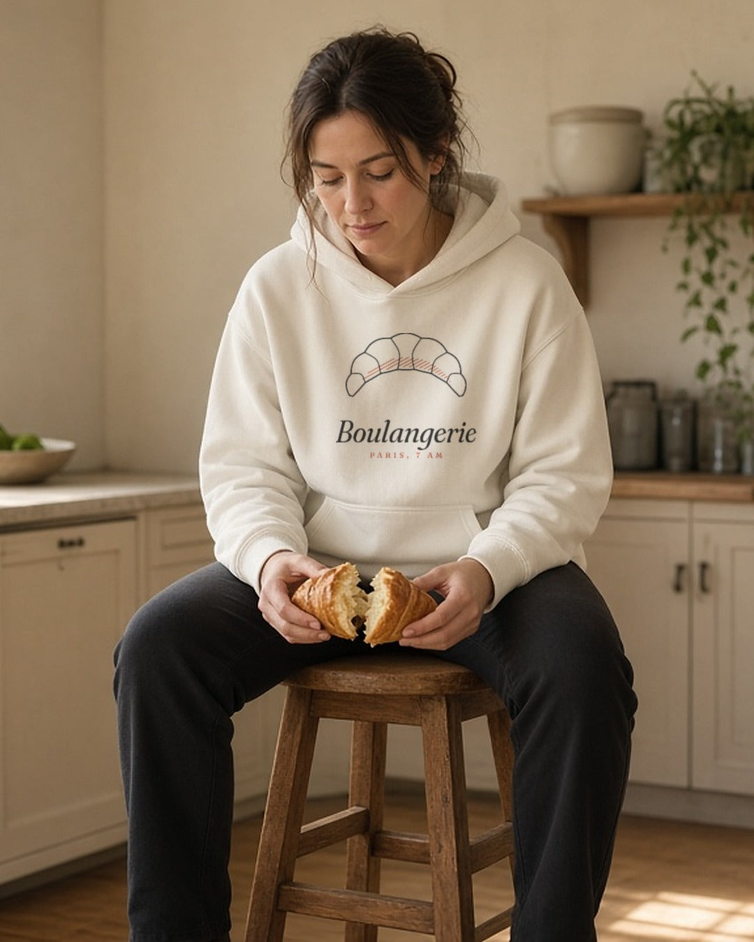 The Boulangerie Hoodie in Bone, a woman on a kitchen stool, knees up inside the hoodie, tearing a croissant