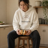 The Boulangerie Hoodie in Bone, a woman on a kitchen stool, knees up inside the hoodie, tearing a croissant