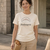 The Boulangerie Tee in Ivory, 7 AM Sharp (7:48): a woman (30s) in the Ivory tee stands outside a small bakery with an empty display window, holding the last croissant in a paper bag like a trophy