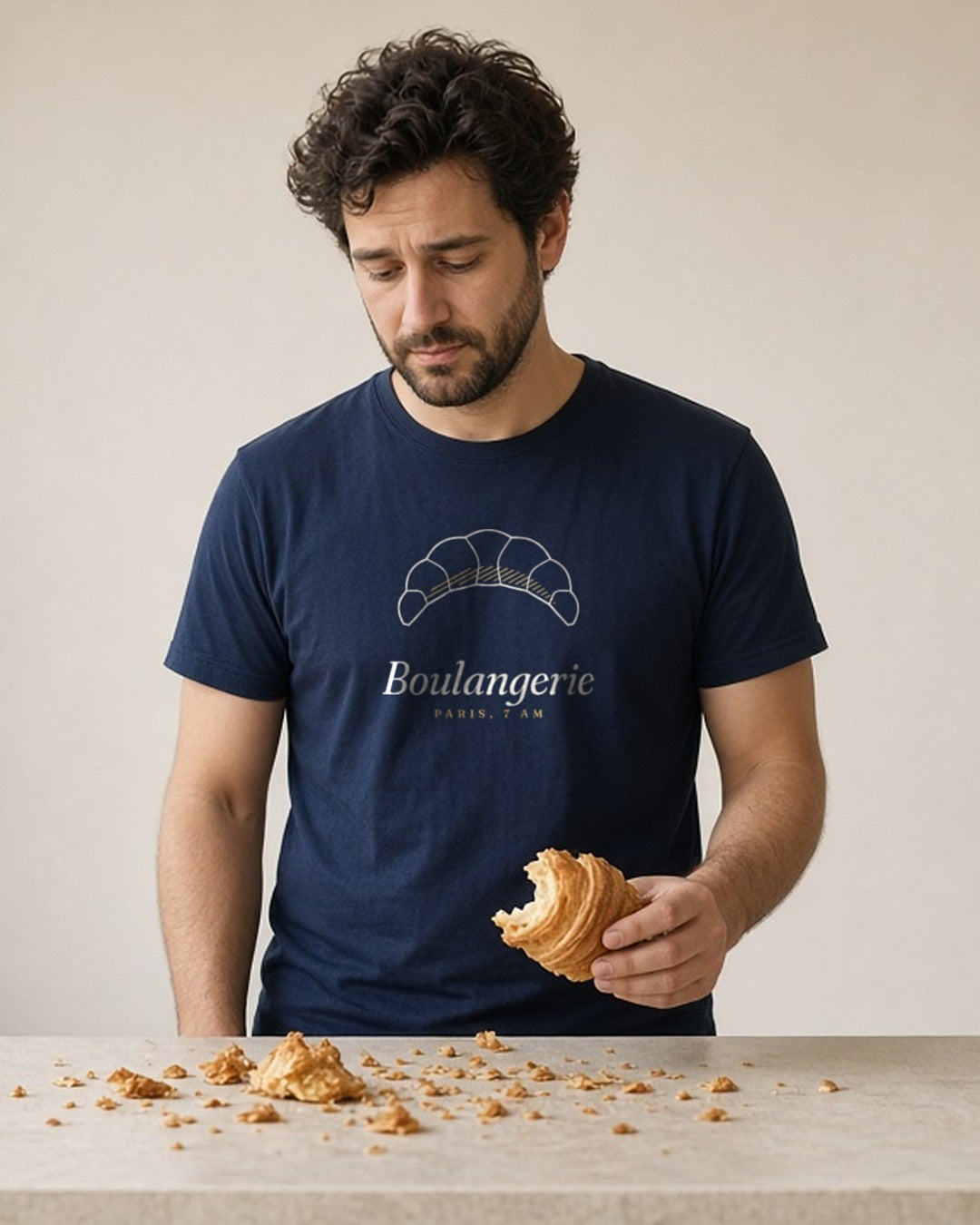 The Boulangerie Tee in True Navy, a man at a café counter, flakes of croissant all over his True Navy tee, looking down at them with mild concern