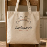 The Boulangerie Tote in Oyster, the tote hung on a chair in a sunny kitchen, a paper bag of pastries beside it