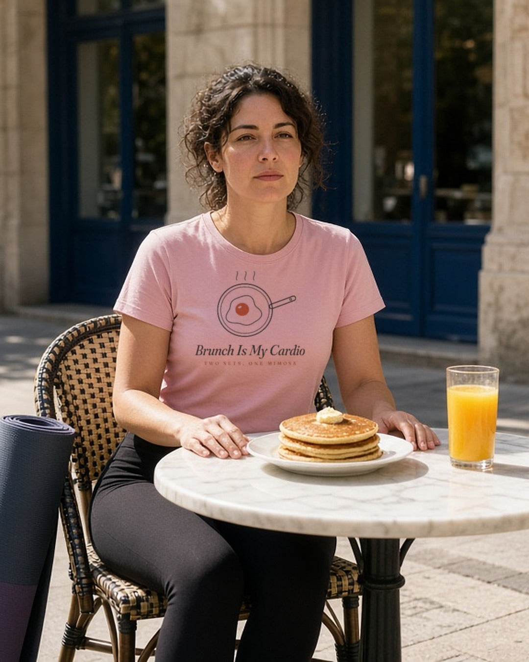 The Brunch Is My Cardio Tee in Blossom, a woman (30s) in workout leggings and the Blossom tee, yoga mat under one arm, seated at a sunny brunch table with pancakes