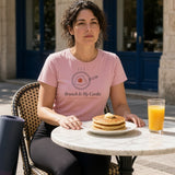 The Brunch Is My Cardio Tee in Blossom, a woman (30s) in workout leggings and the Blossom tee, yoga mat under one arm, seated at a sunny brunch table with pancakes