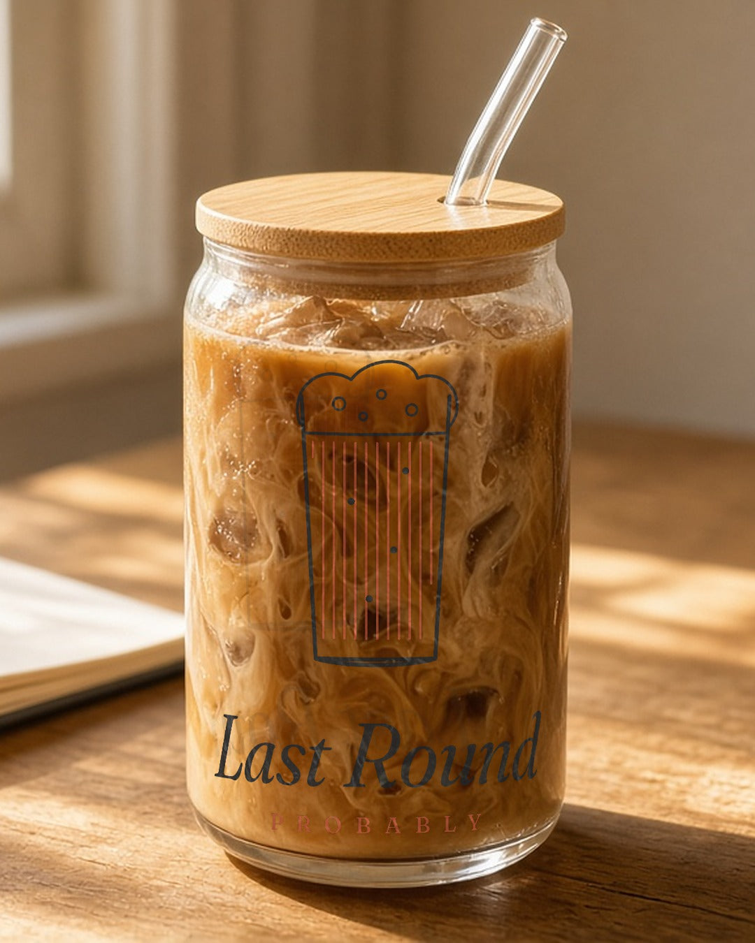 The Last Round Can Glass in Clear 16 oz with lid & straw, can glass with bamboo lid and glass straw full of iced coffee on a sunny wooden desk beside glasses and a notebook