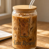 The Last Round Can Glass in Clear 16 oz with lid & straw, can glass with bamboo lid and glass straw full of iced coffee on a sunny wooden desk beside glasses and a notebook