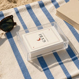 The House Rules Playing Cards, the deck in its acrylic box on a blue-striped towel next to sunglasses and a paperback