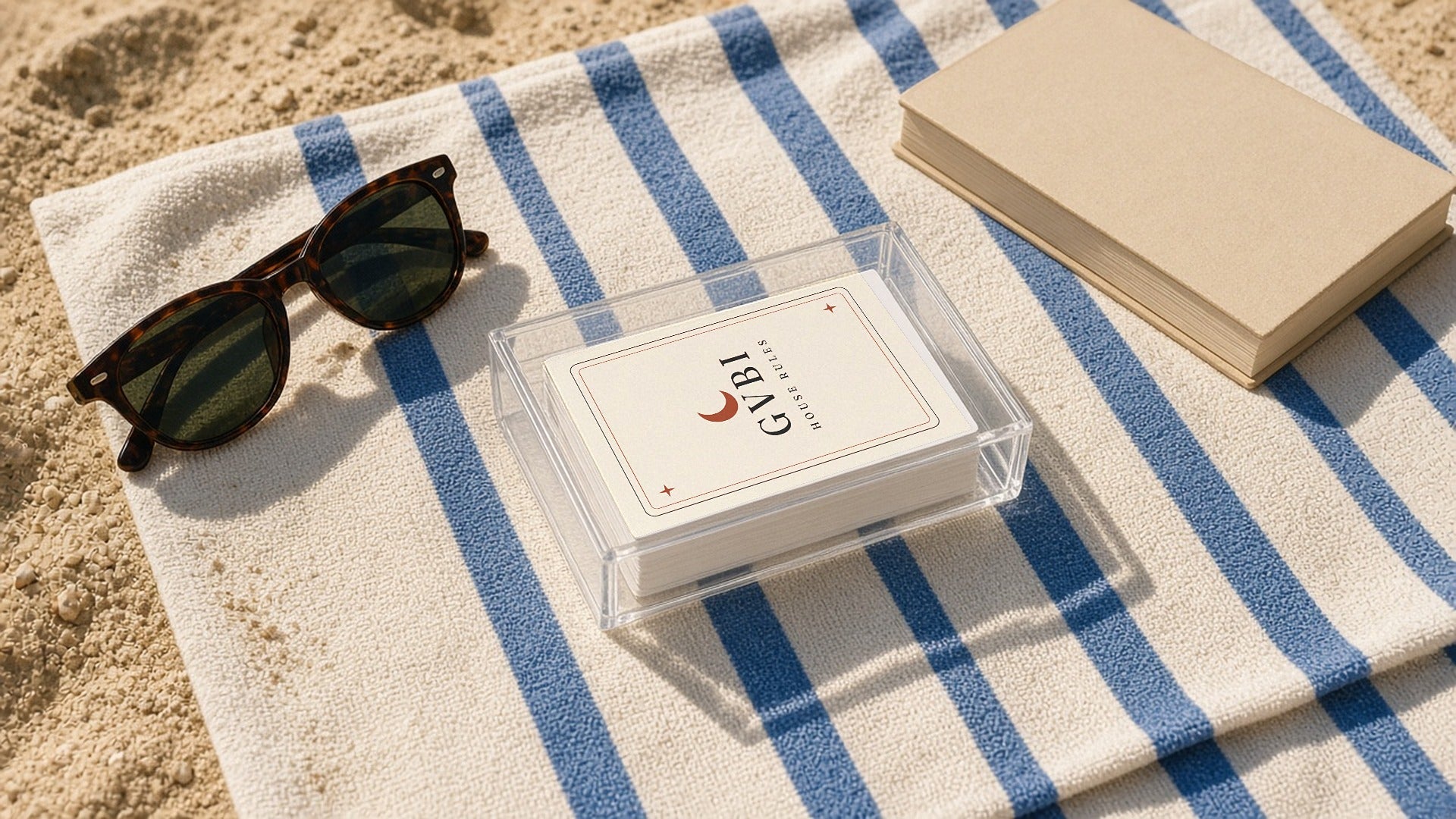 The House Rules Playing Cards, the deck in its acrylic box on a blue-striped towel next to sunglasses and a paperback