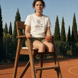 The Clay Season Tee in Ivory, worn by a woman eating a peach on an umpire chair at a clay court