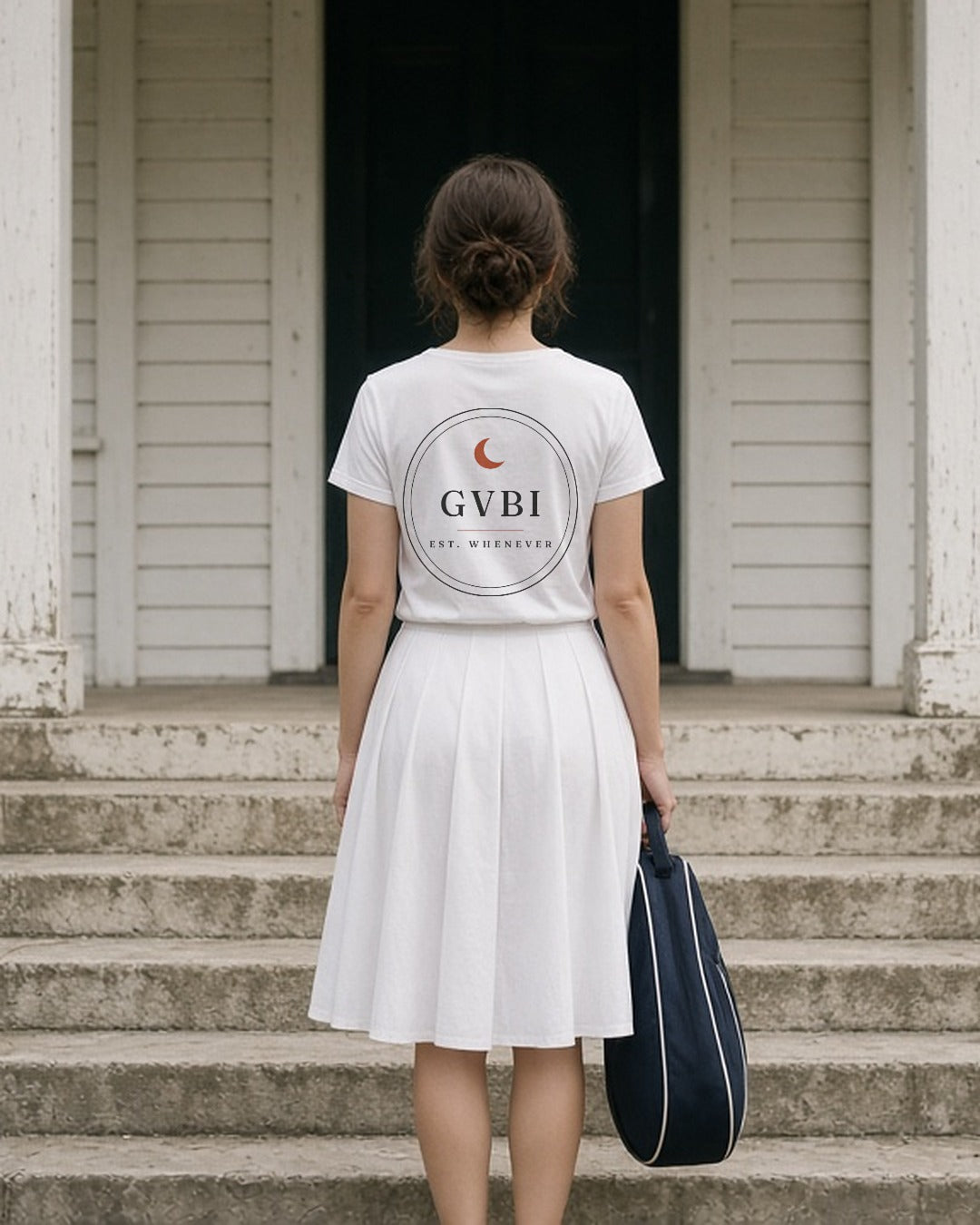 The House Crest Tee in White, woman (late 20s) on white clubhouse steps seen from behind, crest back print, racket bag in hand, soft shade