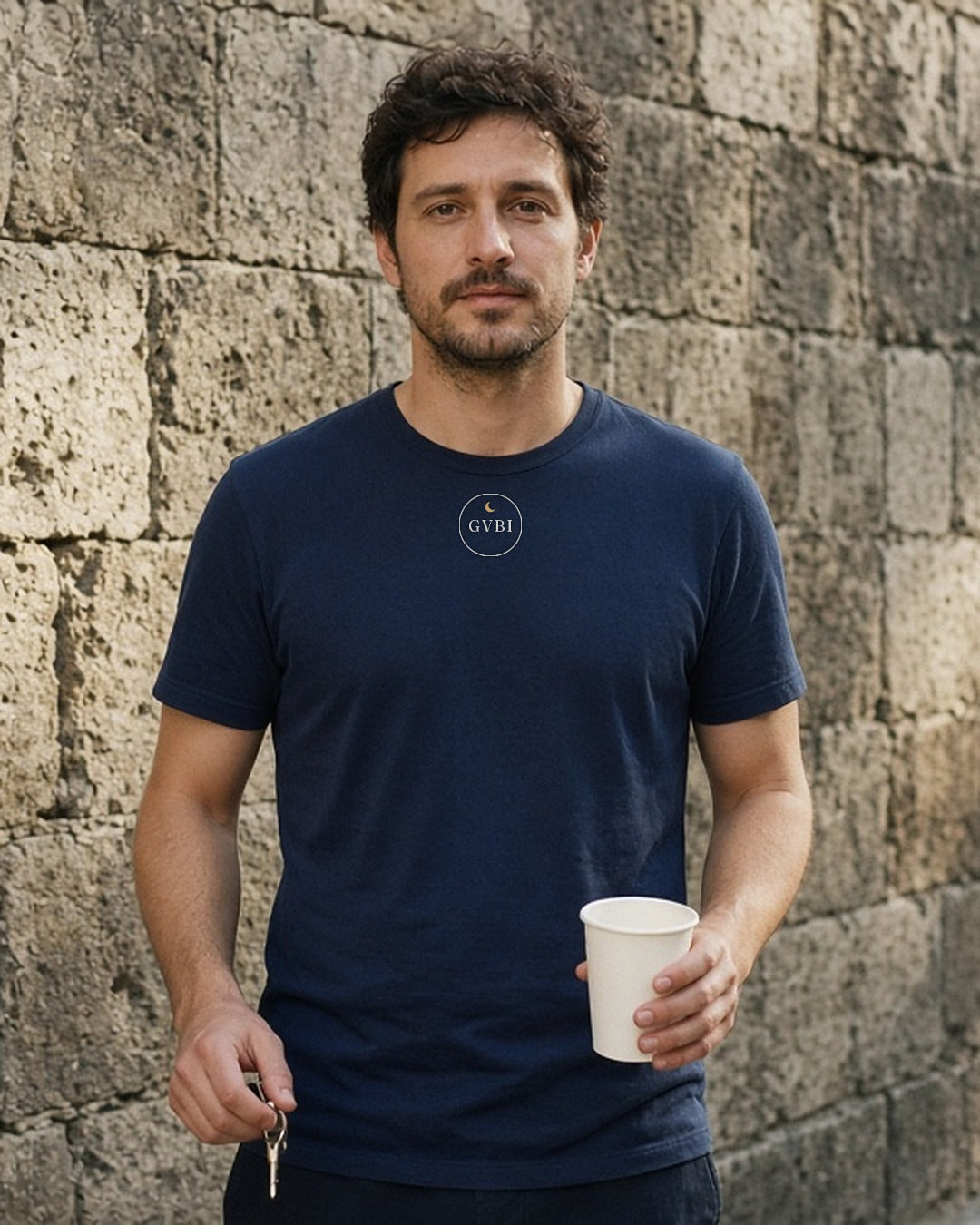 The House Crest Tee in True Navy, man against an old stone wall holding a coffee cup and keys