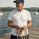 The House Dad Hat in Navy, man (50s) on a wooden dock in the Navy cap, coiling a rope, sailboats behind, hard sun