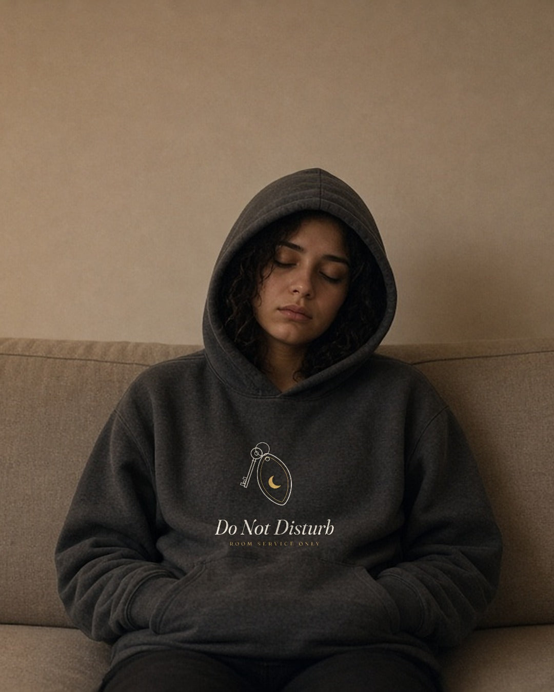 The Do Not Disturb Hoodie in Charcoal Heather, worn hood up by a woman asleep sitting up on a long couch