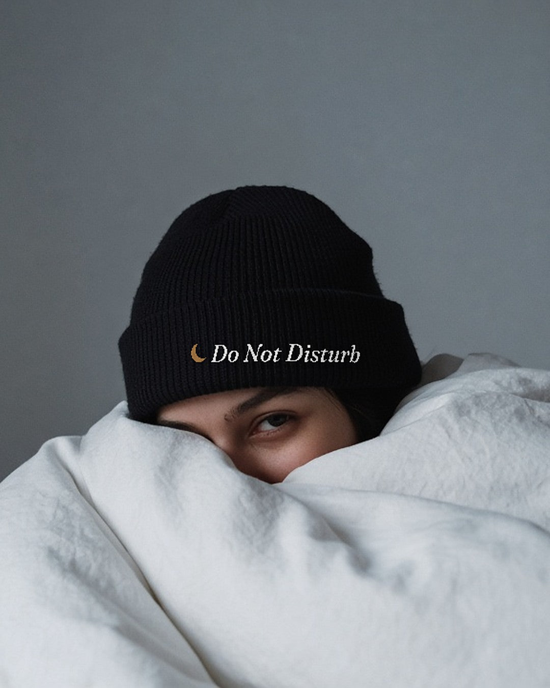 The Do Not Disturb Beanie in Black, worn in bed with the duvet pulled up and an alarm clock nearby