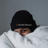 The Do Not Disturb Beanie in Black, worn in bed with the duvet pulled up and an alarm clock nearby