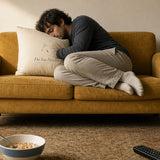 The Do Not Disturb Pillow, hugged by a man curled up asleep on a mustard loveseat