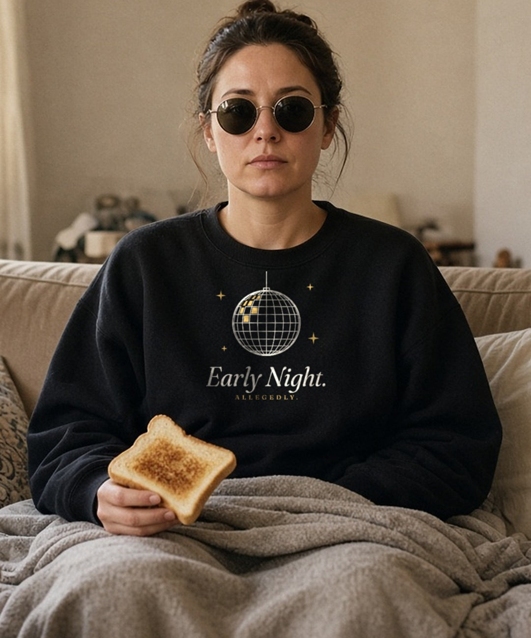 The Early Night Crewneck in Black, a woman on a sofa under a blanket at noon, sunglasses on indoors, toast in hand