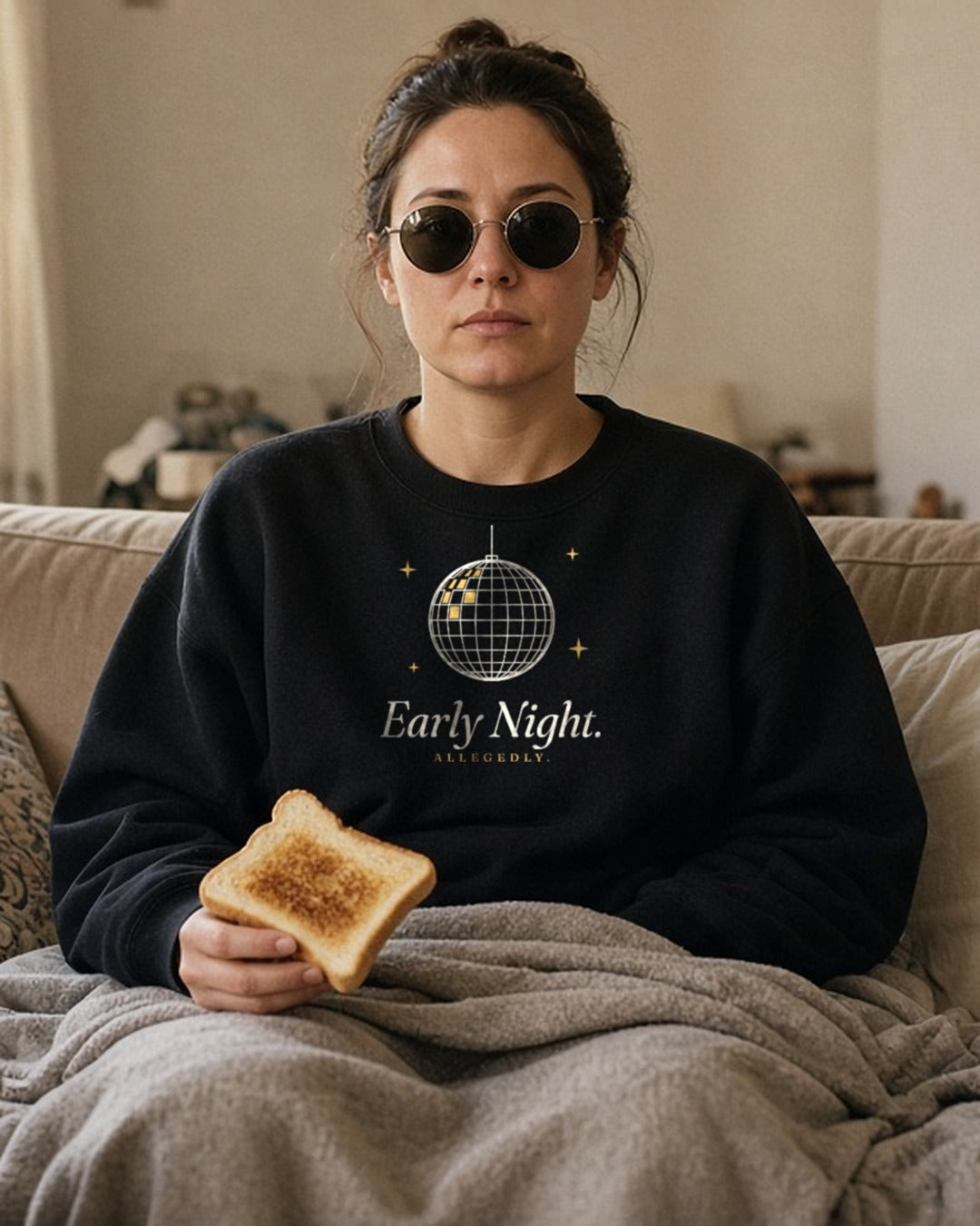 The Early Night Crewneck in Black, a woman on a sofa under a blanket at noon, sunglasses on indoors, toast in hand
