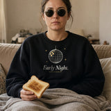 The Early Night Crewneck in Black, a woman on a sofa under a blanket at noon, sunglasses on indoors, toast in hand