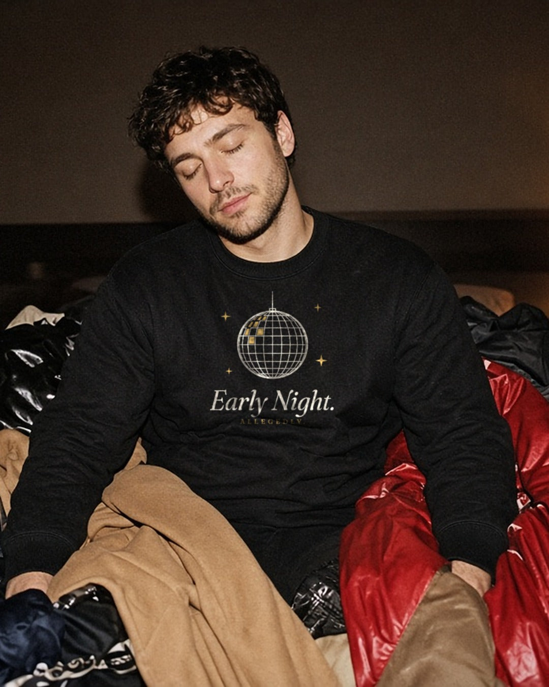 The Early Night Crewneck in Black, young man in the Black crew asleep sitting up on a pile of party coats on a bed (coats run off both sides)