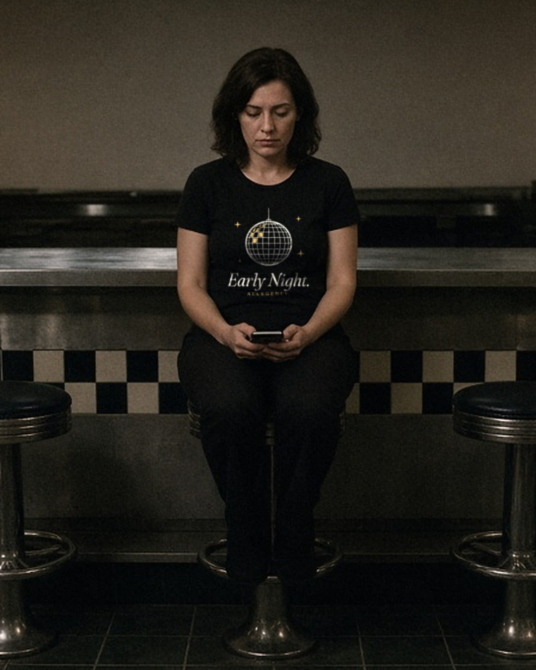 The Early Night Tee in Black, woman in the Black tee alone on a stool at a dim late-night diner counter, phone in her hands