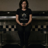 The Early Night Tee in Black, woman in the Black tee alone on a stool at a dim late-night diner counter, phone in her hands