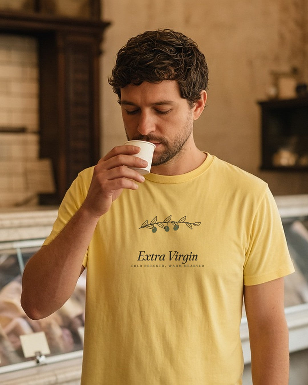 The Extra Virgin Tee in Butter, worn by a man solemnly sniffing a cup of olive oil at a deli counter