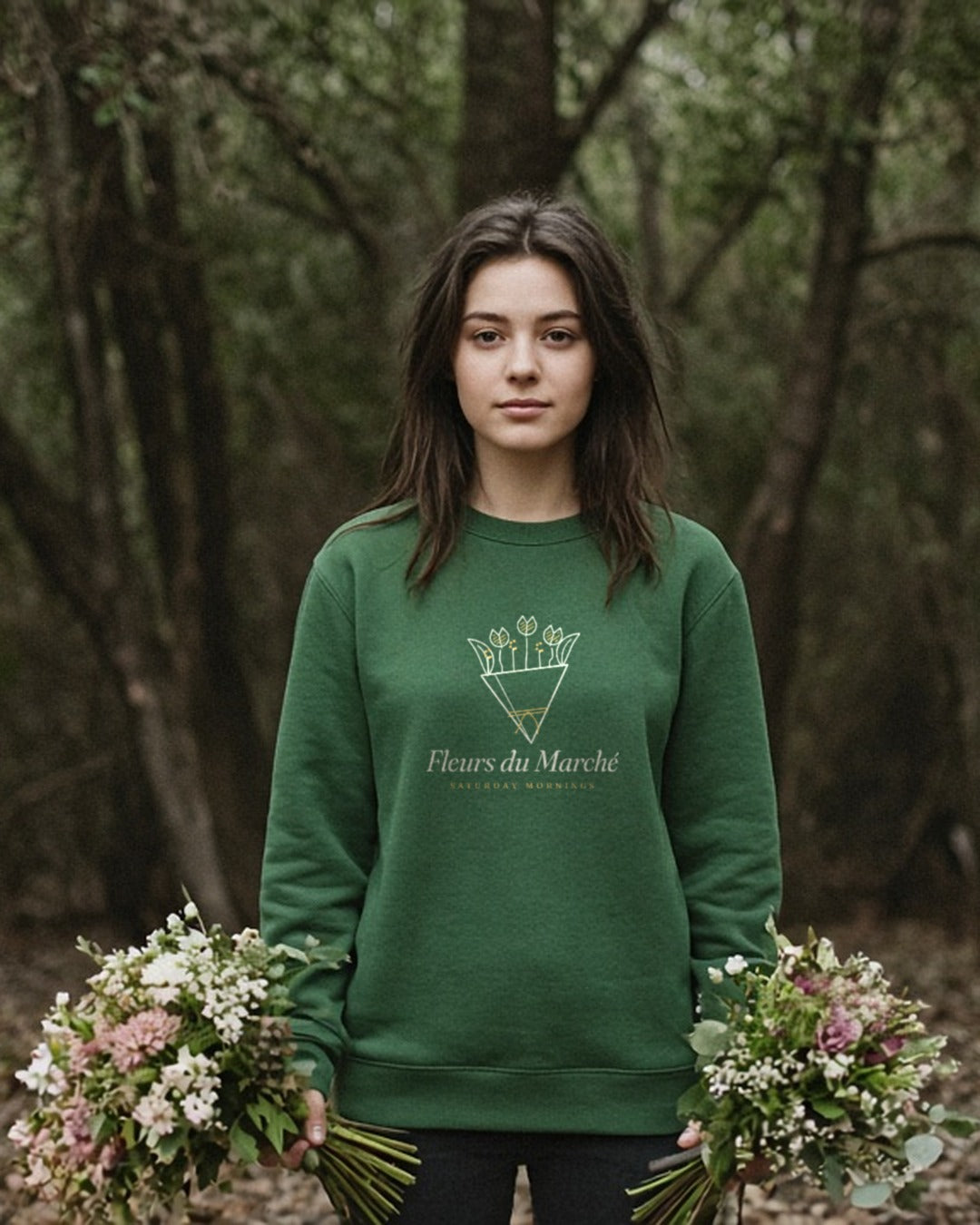 The Fleurs du Marché Crewneck in Forest Green, woman standing in a wooded park, a wildflower bouquet hanging at each side
