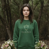 The Fleurs du Marché Crewneck in Forest Green, woman standing in a wooded park, a wildflower bouquet hanging at each side