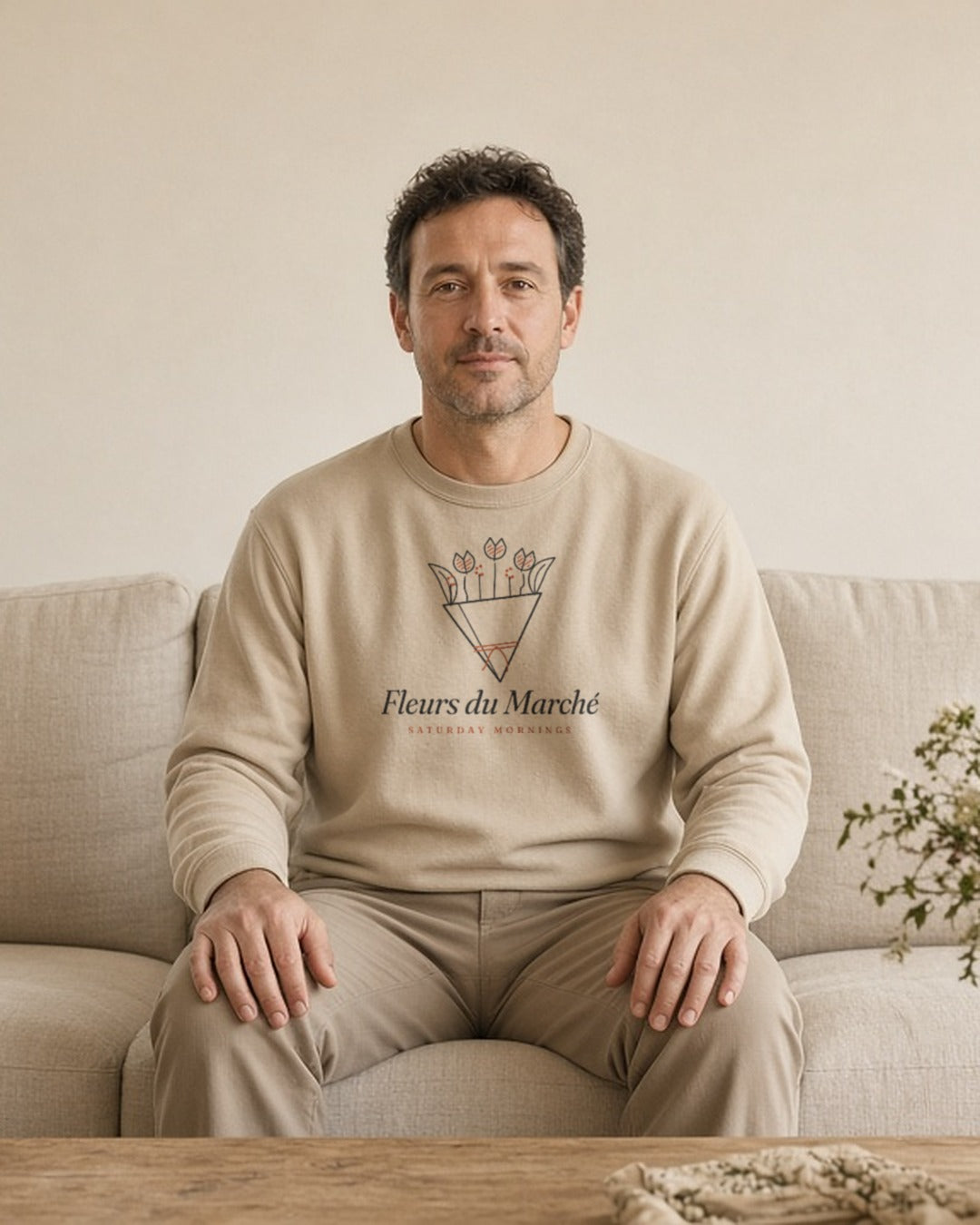 The Fleurs du Marché Crewneck in Sand, a man in Sand on a sofa with a full vase on the coffee table and nowhere to be