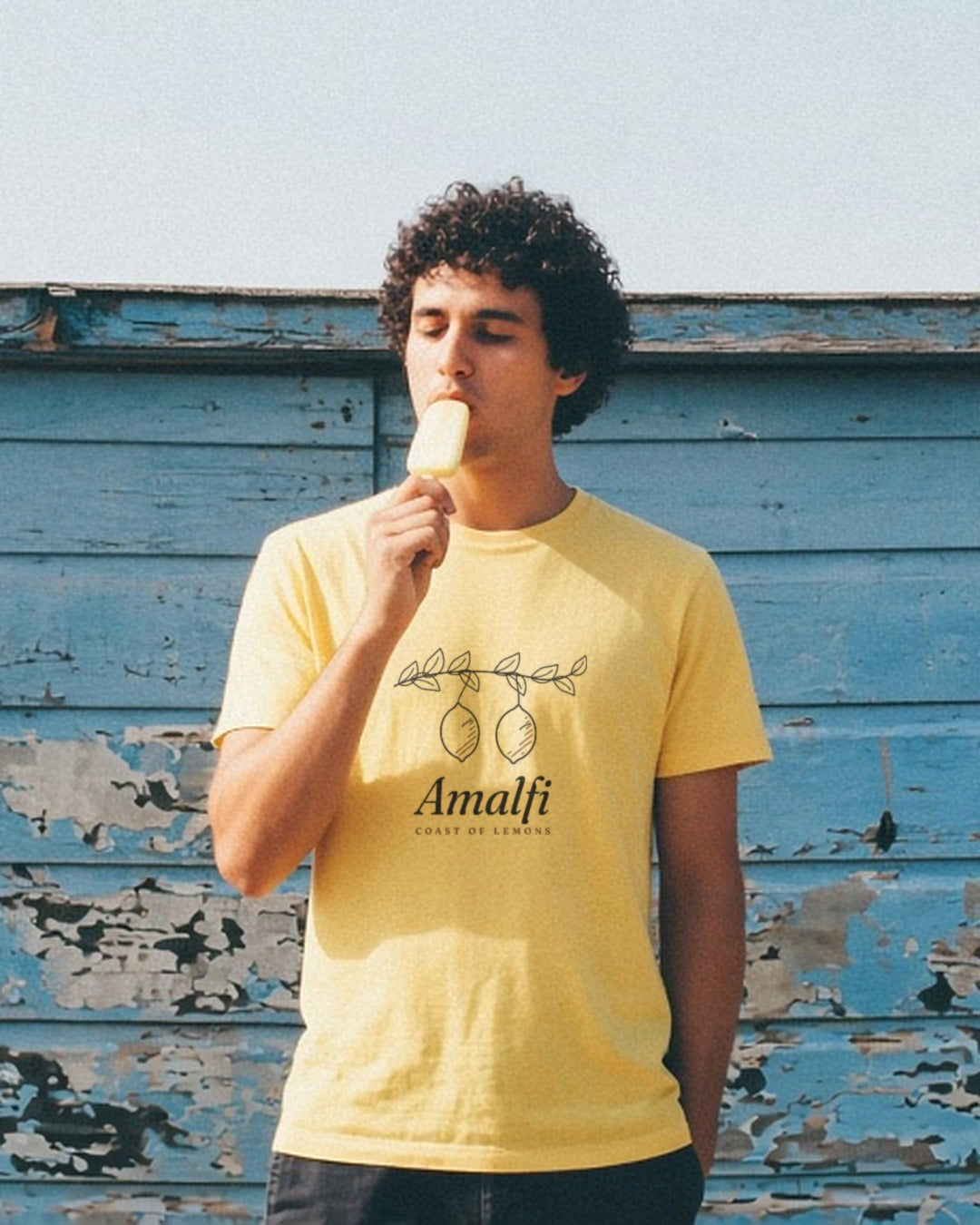 The Amalfi Tee in Butter, eating a popsicle against a peeling blue wall