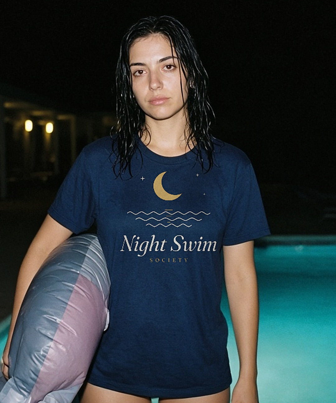 The Night Swim Society Tee in True Navy, by a motel pool at night with an inflatable under one arm