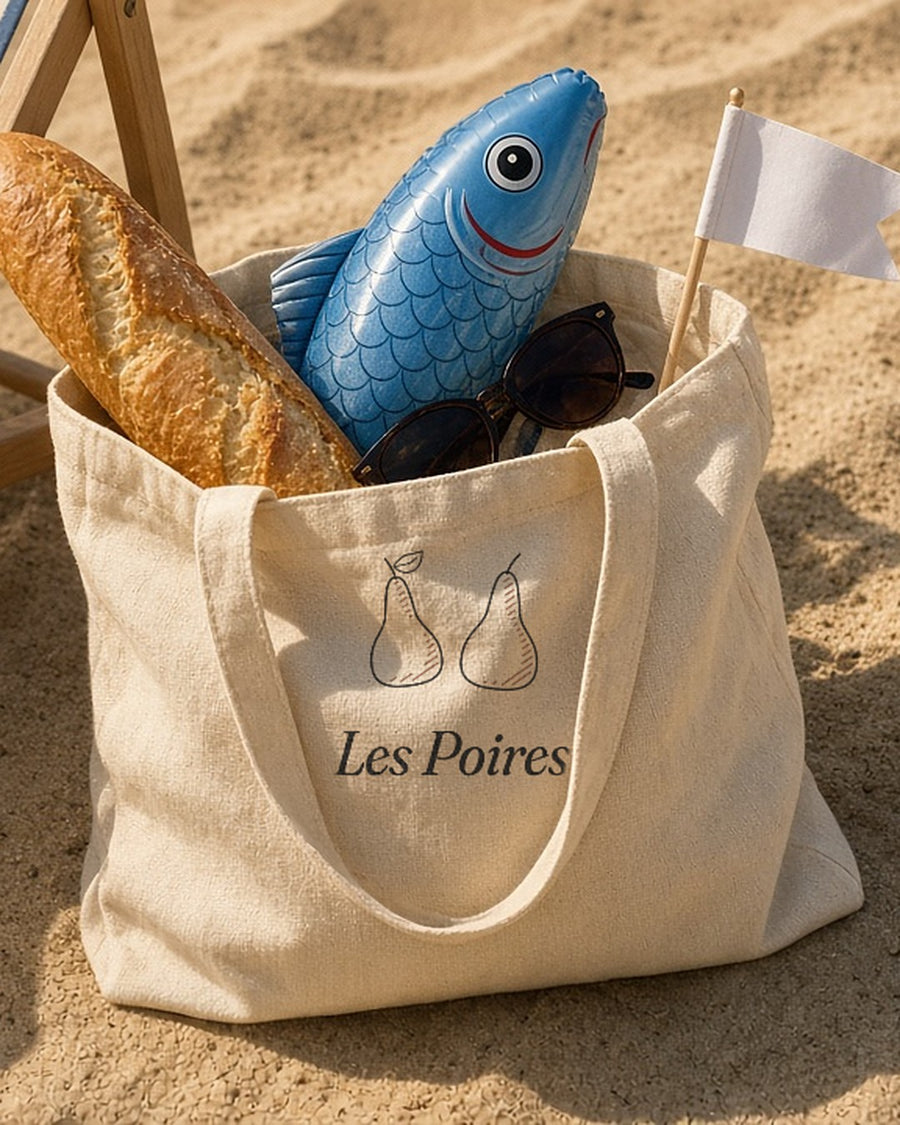 The Les Poires Tote in the sand by a striped beach chair, with a baguette and an inflatable fish