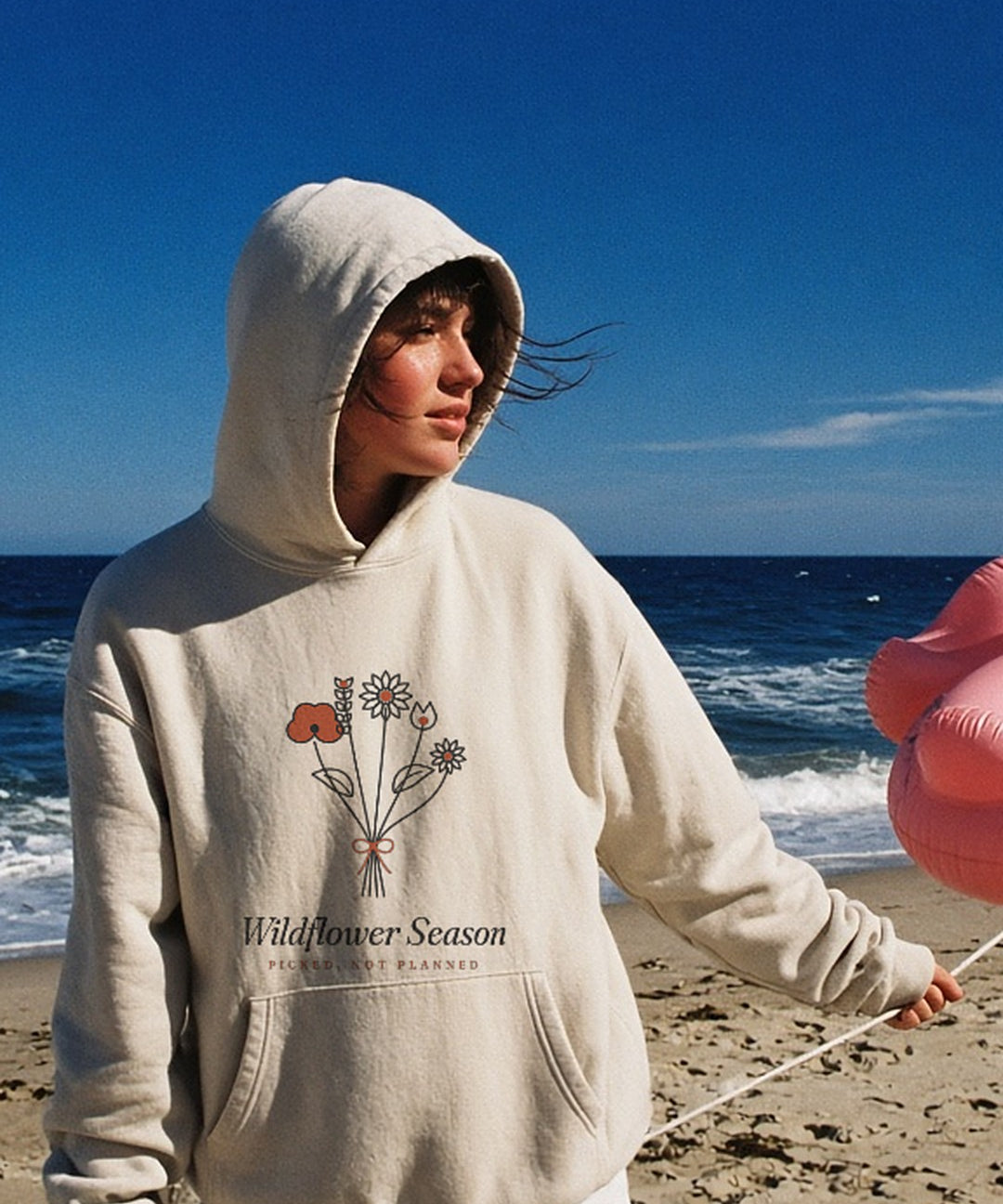 The Wildflower Season Hoodie in Bone, on a windy beach with a flamingo float on a string