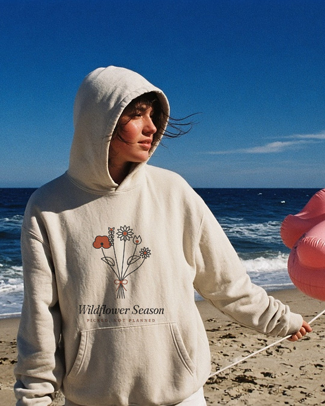 The Wildflower Season Hoodie in Bone, on a windy beach with a flamingo float on a string