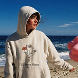 The Wildflower Season Hoodie in Bone, on a windy beach with a flamingo float on a string