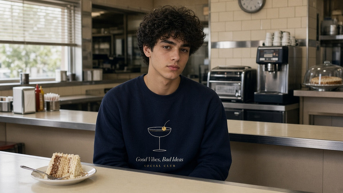 The Social Club Crewneck in Navy, at a diner counter with a slice of cake