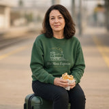 The Hot Mess Express Crewneck in Forest Green, a woman (40s) sitting on her suitcase on an empty platform, eating a sandwich, entirely at peace