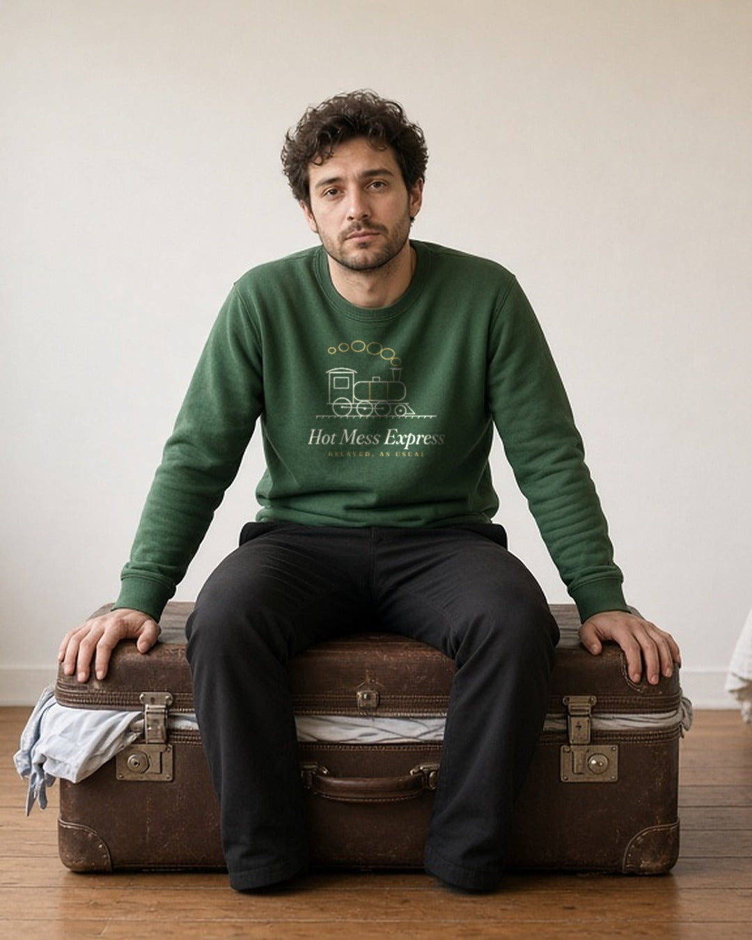 The Hot Mess Express Crewneck in Forest Green, a man sitting on an overstuffed suitcase to close it