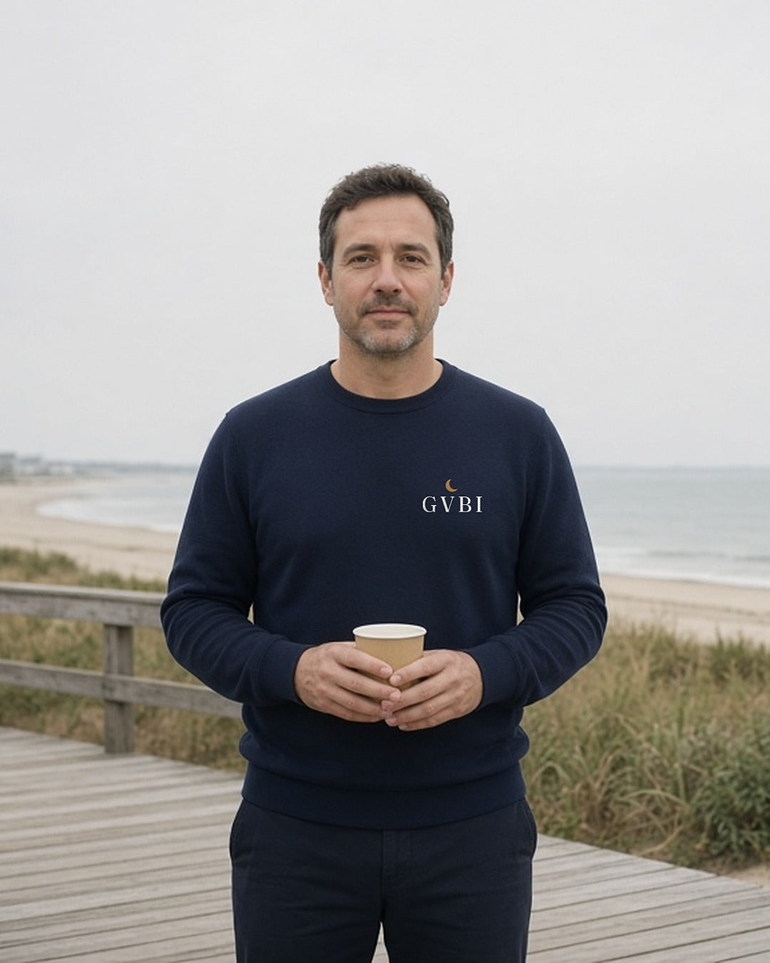 The House Crewneck in Navy, man (40s) on a coastal boardwalk at 7 AM, paper cup in both hands, Navy crewneck, soft overcast light