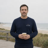 The House Crewneck in Navy, man (40s) on a coastal boardwalk at 7 AM, paper cup in both hands, Navy crewneck, soft overcast light