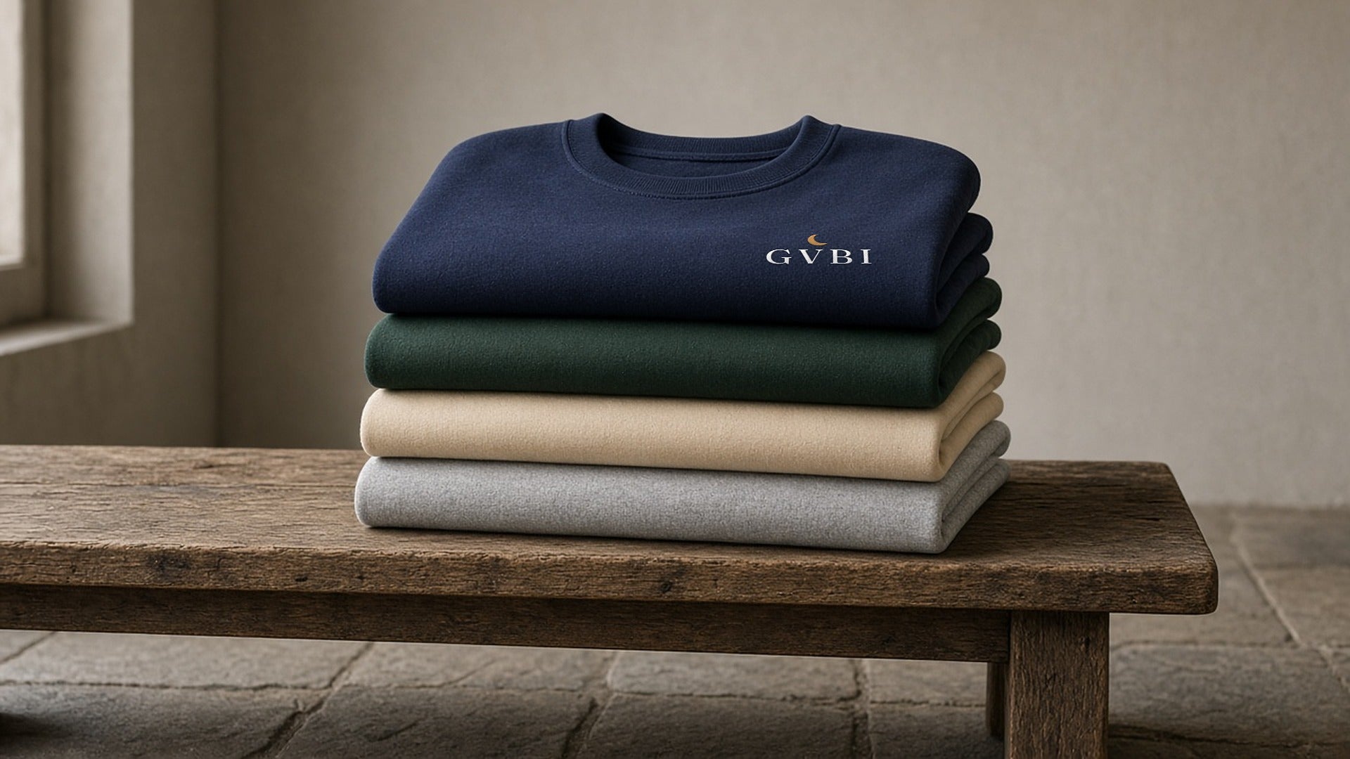 The House Crewneck in Navy, Forest Green, Sand, Ash, all four colours stacked on a wooden bench, soft window light