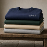 The House Crewneck in Navy, Forest Green, Sand, Ash, all four colours stacked on a wooden bench, soft window light
