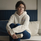 The House Hoodie in Bone, woman (30s) in a window seat with a book in the Bone hoodie, chest embroidery visible, window light