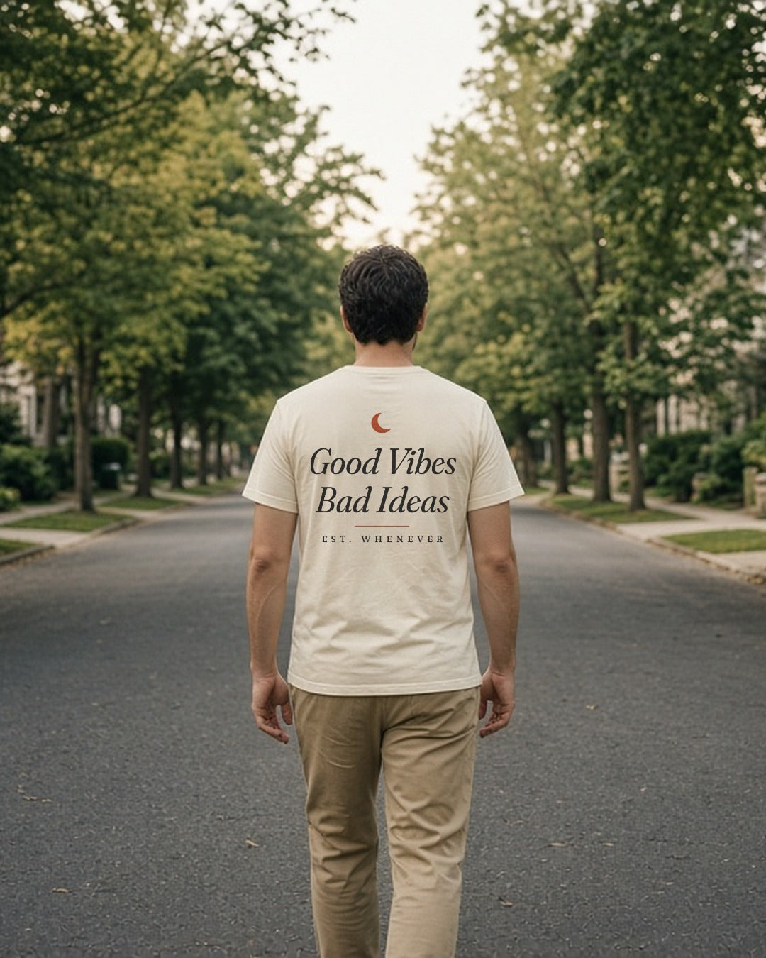 The House Tee in Ivory, man (30s) walks away down a tree-lined street, back print fully readable, soft light