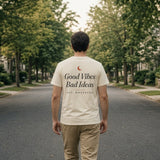 The House Tee in Ivory, man (30s) walks away down a tree-lined street, back print fully readable, soft light