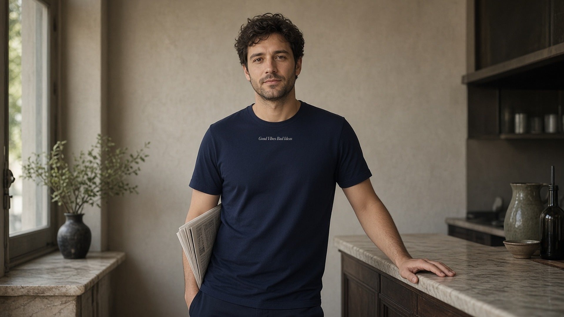 The House Tee in True Navy, same-type man at a café counter, small wordmark on chest, newspaper under his arm